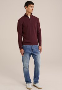 Pulover cu guler tip fermoar în nuanță burgundy, din material tricotat, asortat cu blugi albaștri deschiși și adidași albi cu talpă bej. Design simplu, croială standard.