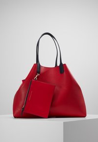 Czerwona skórzana torba tote o luźnym fasonie, z czarnymi uchwytami i odpinaną kieszonką na zamek w dopasowanej czerwieni. Gładka tekstura.