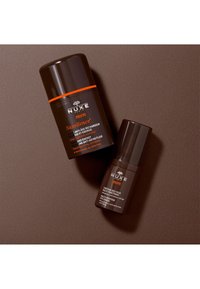 NUXE herrhudvårdsprodukter, mörkbrown flaskor med orange detaljer, en stor vätskebehållare och en mindre ögonkräm, släta ytor.