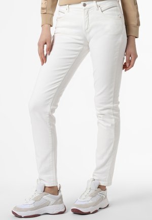Personne portant un jean blanc ajusté et des baskets blanc beige sur un fond uni, montrée de la taille aux pieds.