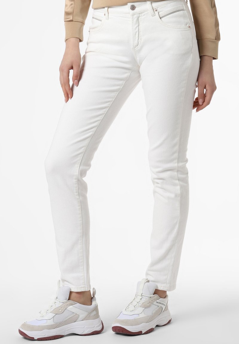 Personne portant un jean blanc ajusté et des baskets blanc beige sur un fond uni, montrée de la taille aux pieds.