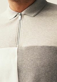 Polo gris à fermeture éclair avec un panneau texturé crème. Tissu doux et lisse avec un col classique et des blocs de couleurs contrastantes.