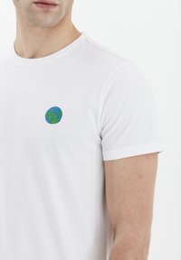 Hvit bomulls t-skjorte med rund hals, med en liten brodert globusdesign i blått og grønt på venstre brystområde.