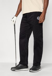 Pantalon en velours côtelé noir à rayures verticales, avec un logo sur la poche avant. Porté avec une chemise beige et des chaussures de golf noires.