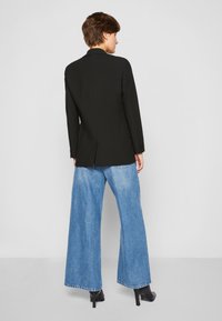 Blazer nero su misura con una fessura centrale sul retro, abbinato a jeans larghi azzurri. Il modello indossa scarpe con tacchi alti neri.