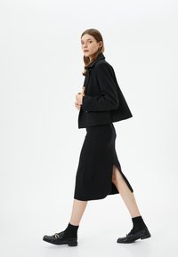 Zwarte cropped jasje van textiel, gecombineerd met een zwarte rok tot halverwege de knie met een zijsplit. Zwarte schoenen met gespen maken de look compleet.