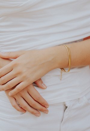 Braccialetti di metallo dorato con una finitura liscia, indossati su una mano con unghie naturali, appoggiata su un tessuto bianco e strutturato.