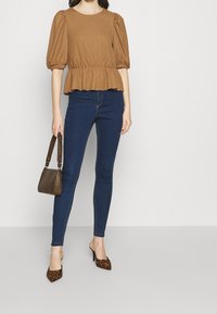 Top marrón con mangas abullonadas y dobladillo en peplum, combinado con jeans ajustados azul oscuro y mules de estampado de leopardo; sosteniendo un pequeño bolso de mano con patrón.