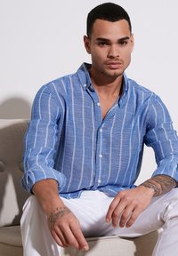 Camicia a maniche lunghe a righe blu e bianche con colletto abbottonato, realizzata in tessuto texturizzato. Modello seduto, indossa pantaloni bianchi.