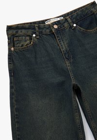 Jeans holgados de mezclilla en azul oscuro, con cinco bolsillos, lazos para cinturón y herrajes de cobre, con un acabado desgastado y ligeras marcas de desgaste.