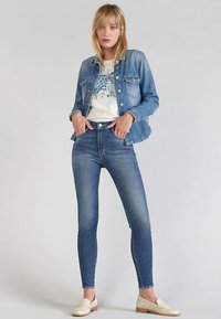 Smala blå jeans med hög midja, kombinerade med en ljus denimjacka och en vit t-shirt med tryck, kompletterade med beige loafers.