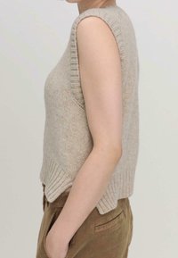 Světle béžový pletený bezrukávový svetr s žebrovaným výstřihem a spodním lemem, ve volném střihu a texturovaném povrchu. V kombinaci s hnědými kordovými kalhotami.