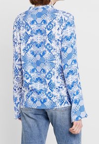 Camicia a maniche lunghe con motivi floreali blu e bianchi, dotata di colletto e maniche svasate, realizzata in tessuto leggero.