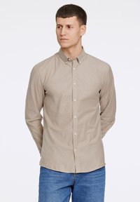 Camicia beige a maniche lunghe con colletto, caratterizzata da una texture liscia e una vestibilità regolare, abbinata a jeans blu.
