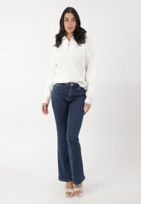 Pull en tricot blanc avec col zippé mi-haut, associé à un jean évasé bleu foncé. Le mannequin porte des talons à lanières de couleur claire sur un fond uni.