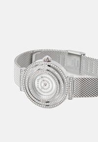 Srebrny zegarek na siateczkowym pasku z okrągłą tarczą z wzorami spiralnymi i okrągłymi akcentami z cyrkoniami. Przezroczysta tarcza z srebrnymi wskazówkami i brandingiem.