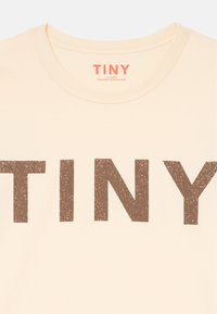 T-shirt couleur crème mettant en avant le mot "TINY" en police marron large et texturée. Encolure ronde, manches courtes, tissu doux.