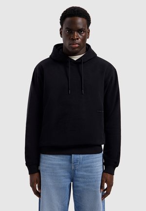 Jonge man met een serieuze blik, gekleed in een zwarte hoodie en lichtblauwe spijkerbroek, staand tegen een effen grijze achtergrond.