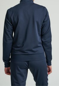 Meriino sininen urheilupuku takki, jossa on korkea kaulus, sileä pinta ja ribbireunat. Sisältää valkoiset raidat hartioiden varrella ja istuvan muotoilun.