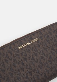 Portefeuille en cuir marron avec une surface texturée, un logo doré "MICHAEL KORS" et une fermeture éclair avec de petits accents métalliques.