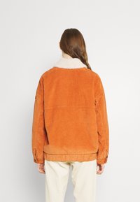 Veste en velours côtelé orange brûlé avec un col sherpa doux couleur crème, dotée de manches longues et d'une coupe décontractée.