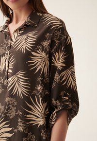 Chemise noire avec un imprimé botanique de feuilles et de fleurs crème, dotée d'un col et de manches retroussées, fabriquée à partir d'un tissu léger et lisse.