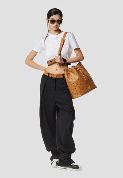Femme portant un tee-shirt blanc court et un pantalon noir ample, portant de grandes lunettes de soleil, avec un sac seau de créateur couleur tan sur l'épaule.