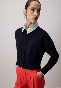 Cardigan bleu marine à boutonnière et ourlet côtelé, porté par-dessus une chemise à col ornée de broderies colorées. Associé à un pantalon rouge vif.