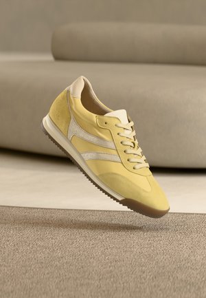 Baskets jaunes avec des accents en daim beige, des lacets blancs et une semelle en caoutchouc flottant au-dessus d'un tapis beige texturé dans une pièce minimaliste.