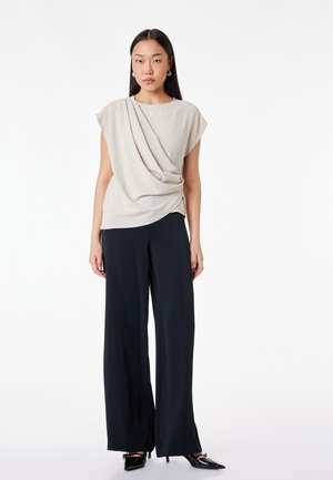 Mujer con una blusa drapeada de manga corta color beige claro, pantalones negros de pierna ancha y tacones negros puntiagudos, de pie con expresión neutral.