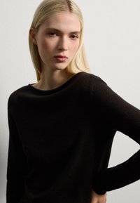 Top nero a maniche lunghe realizzato in materiale morbido, con scollatura rilassata e maniche aderenti, mostrato su uno sfondo bianco semplice.