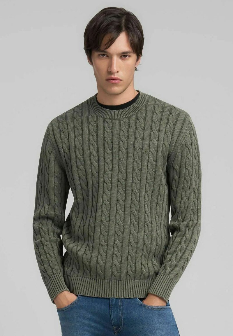 Maglione verde a trecce con scollo e polsini a coste, caratterizzato da motivi texture. Indossato sopra una camicia nera e abbinato a jeans blu.