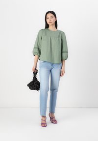 Blusa verde texture con maniche a sbuffo, abbinata a jeans azzurri chiari. Tiene una borsa nera con motivo e indossa tacchi alti viola con cinturini.