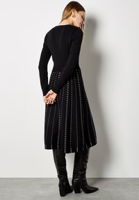 Vestido negro de punto con mangas largas, textura acanalada en el cuerpo y una falda acampanada con rayas blancas verticales. Botas negras hasta la rodilla.