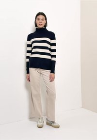 Pull à col roulé rayé bleu marine et crème, associé à un pantalon ample beige et des baskets avec des accents bleus et gris.