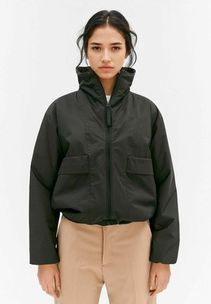 Kvinde iført sort lynlåsjakke med høj krave og tanfarvede bukser, stående mod en ensfarvet hvid baggrund.