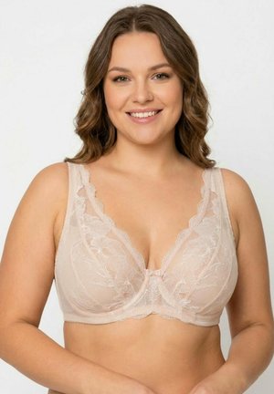 Uśmiechnięta kobieta z falującymi brązowymi włosami, ubrana w beżowy koronkowy biustonosz z fiszbinami na jednolitym jasnym tle.