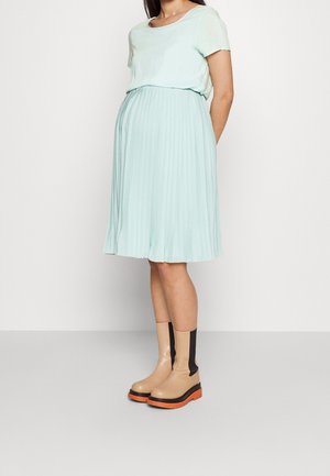 Hellgrünes, plissiertes Kleid mit kurzen Ärmeln, kombiniert mit beigen Stiefeletten mit schwarzem Akzent und oranger Sohle.