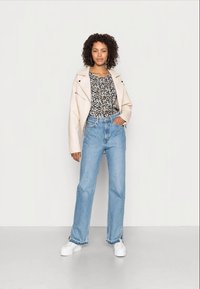 Jaqueta de couro bege sobre uma camisa floral, combinada com jeans de cintura alta em azul claro e tênis brancos. Cabelo cacheado visível.