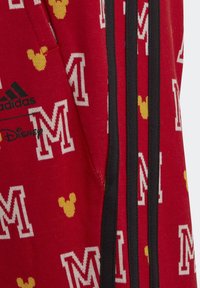 Pantalones de chándal rojos de Adidas con un patrón de íconos amarillos de Mickey Mouse y letras 'M' blancas, con franjas laterales negras y acentos de logo.
