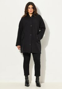 Cappotto nero con cappuccio, dal taglio morbido, con chiusura a grandi bottoni e due tasche laterali. Abbinato a pantaloni neri con texture e stivaletti alla caviglia.