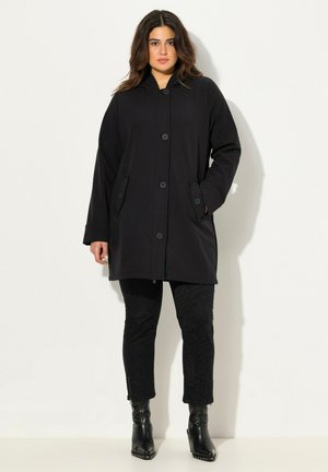 Cappotto nero con cappuccio, dal taglio morbido, con chiusura a grandi bottoni e due tasche laterali. Abbinato a pantaloni neri con texture e stivaletti alla caviglia.