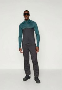 Ensemble de vêtements de sport pour hommes comprenant une veste zippée teal et noire à manches longues et col montant, assortie de pantalons noirs ajustés et de baskets noires.