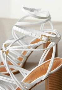 Hvide strappy højhælede sandaler med flettet detaljer, guldfittings og en stablet træhæl. Glat læder og strukturerede stropper.