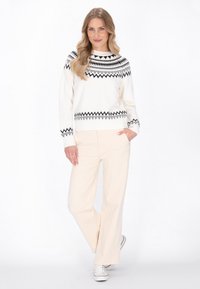 Maglione bianco con motivi geometrici neri, scollatura rotonda e maniche lunghe, abbinato a pantaloni larghi beige chiaro e sneakers bianche.