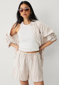 Camiseta sin mangas blanca combinada con una camisa a rayas en tonos marrón claro y blanco y pantalones cortos a juego. Gafas de sol con un suave acabado de lentes degradados.