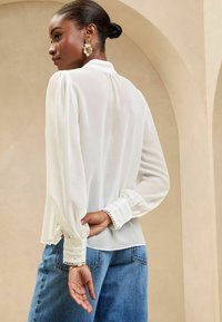Camicia bianca con maniche a sbuffo e dettagli in pizzo al colletto e ai polsini, abbinata a jeans in denim blu, presentata su uno sfondo neutro.