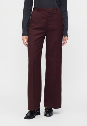 Personne portant un pantalon large bordeaux foncé, des mocassins noirs polis et un haut à manches longues gris foncé, debout devant un fond uni.