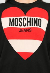 Svart genser med et stort rødt og hvitt hjerte-design. Sentrert tekst med "MOSCHINO JEANS" i fete svarte bokstaver.
