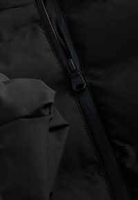 Detail einer schwarzen Steppjacke mit einem glatten Reißverschluss und gefaltetem Stoff. Das Material ist leicht und hat eine glänzende Oberfläche.